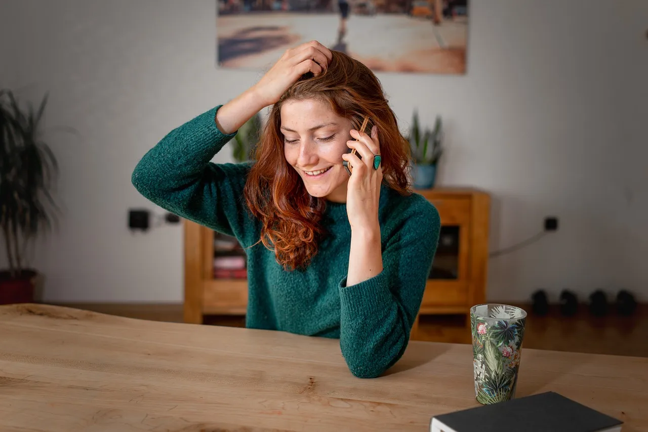 #FotoDescrição: Mulher sorrindo enquanto fala ao celular, sentada à mesa com uma caneca e objetos de decoração ao fundo.