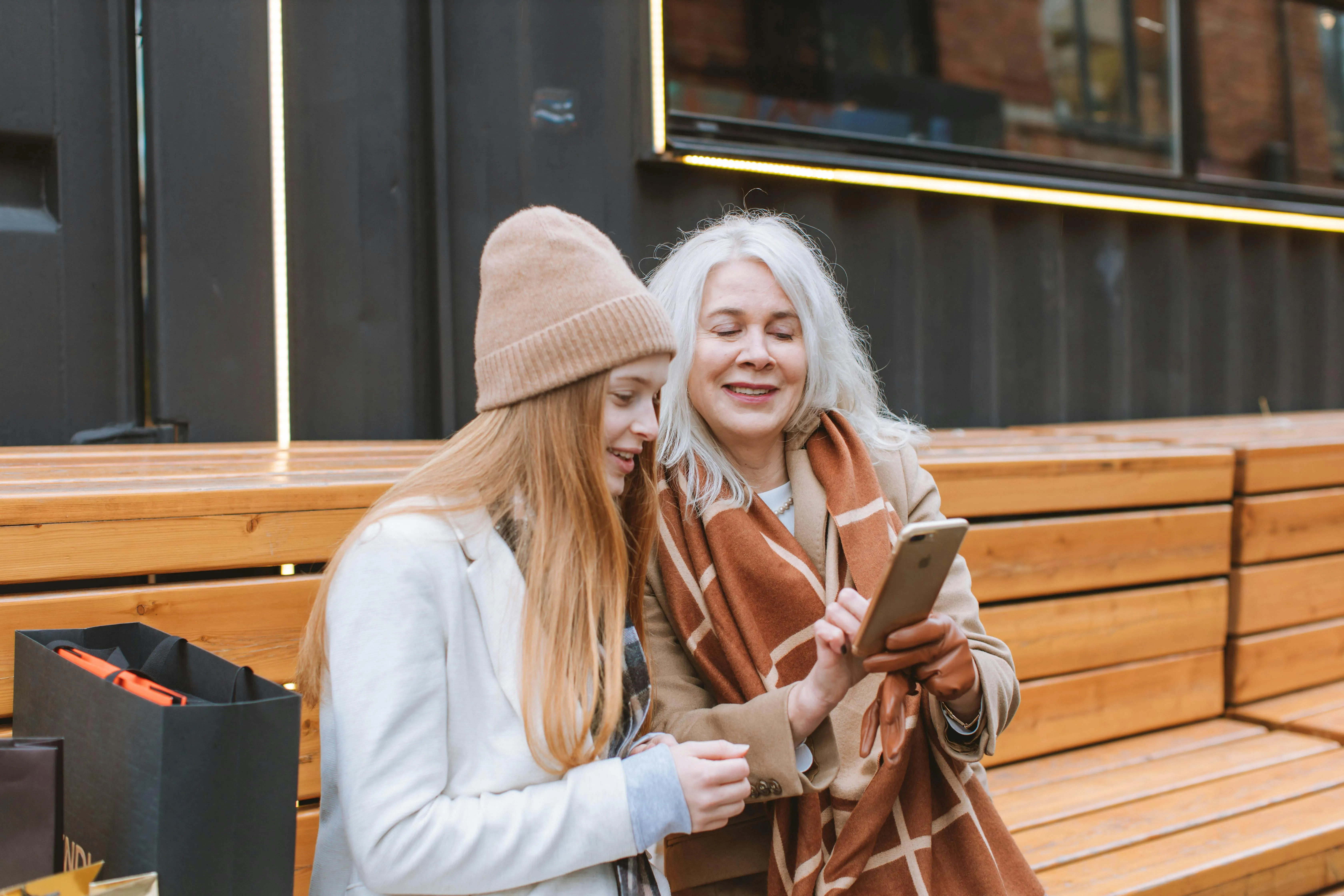 #FotoDescrição: Duas mulheres sentadas em um banco de madeira ao ar livre, sorrindo enquanto olham para a tela de um smartphone.