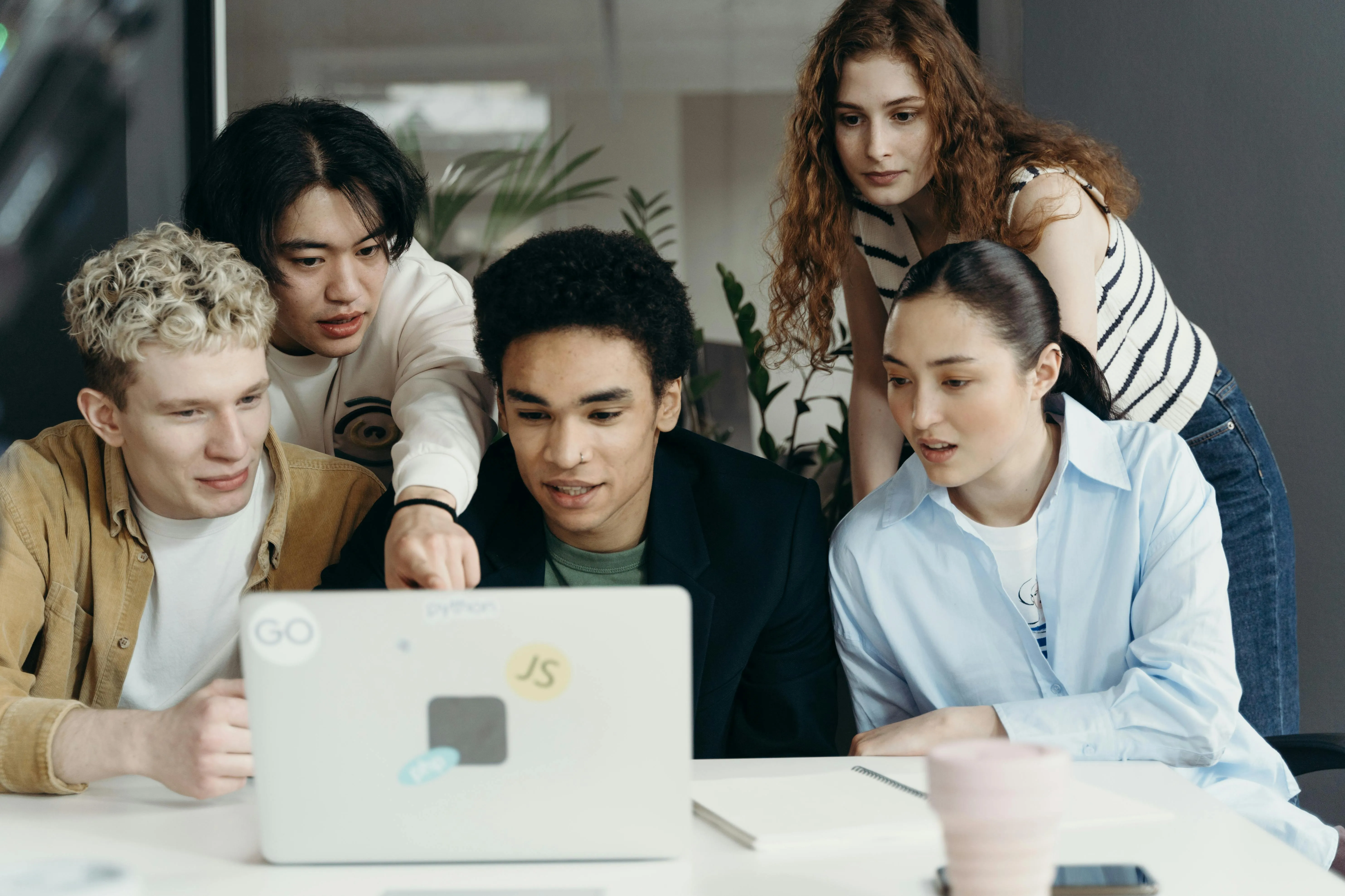 #FotoDescrição: Cinco jovens reunidos em torno de um notebook sobre a mesa, observando algo na tela com atenção.