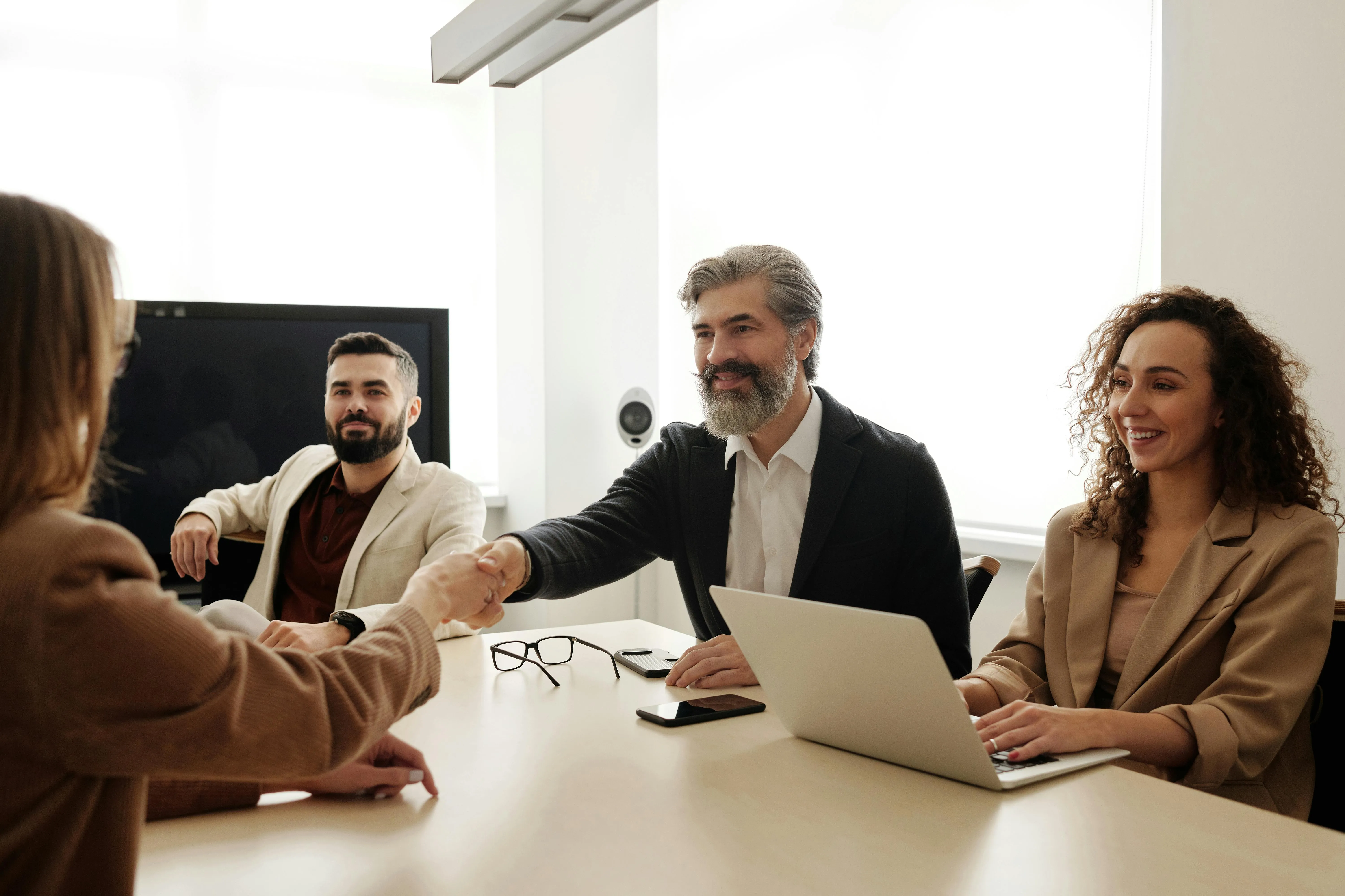 #FotoDescrição: Reunião de negócios com pessoas sorrindo, enquanto duas delas apertam as mãos em sinal de acordo.uanto duas delas apertam as mãos em sinal de acordo.