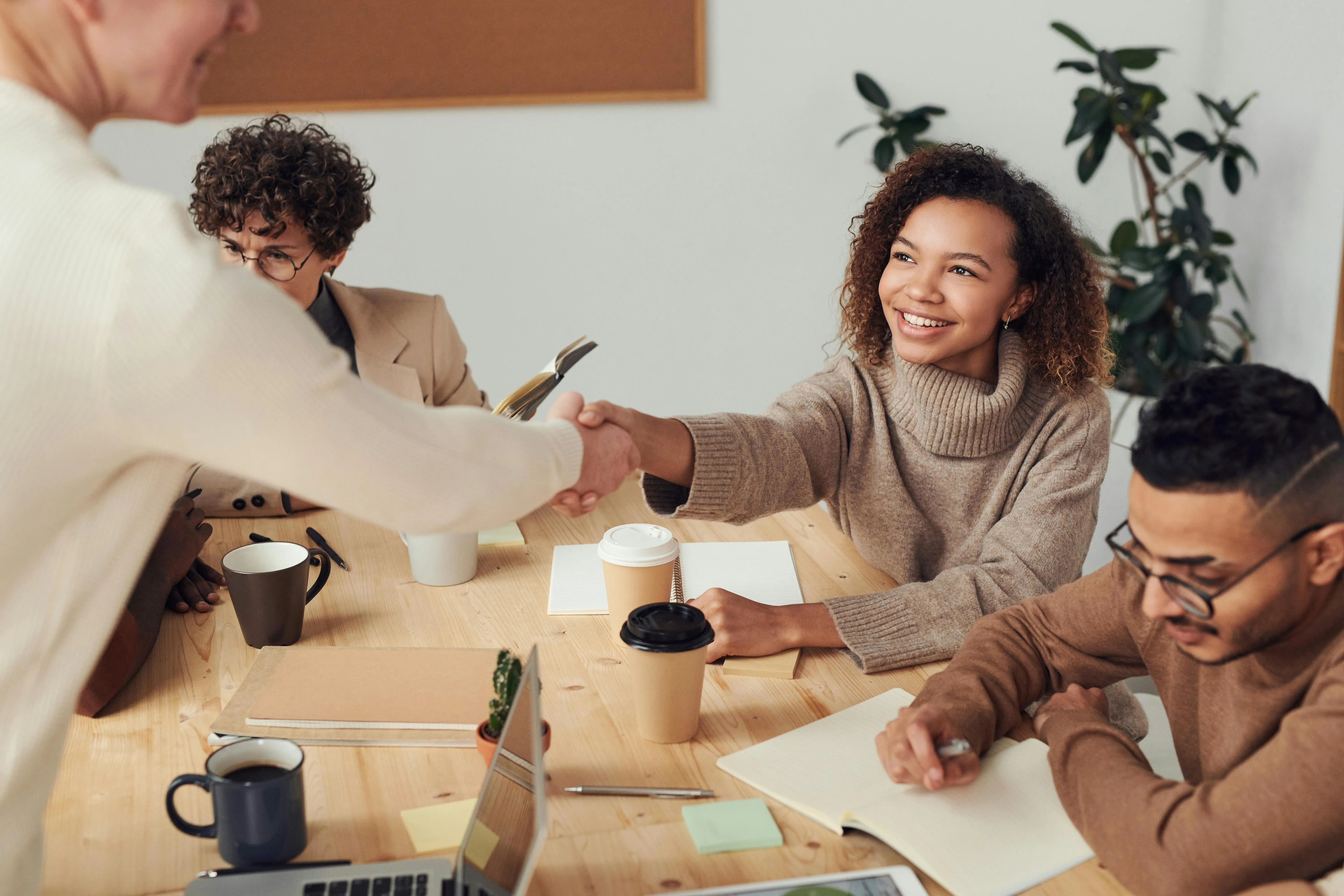 #FotoDescrição Grupo reunido em uma mesa de reunião, onde uma mulher sorridente aperta a mão de outra pessoa, simbolizando acordo, parceria ou fechamento de negócio com clima amigável e colaborativo.