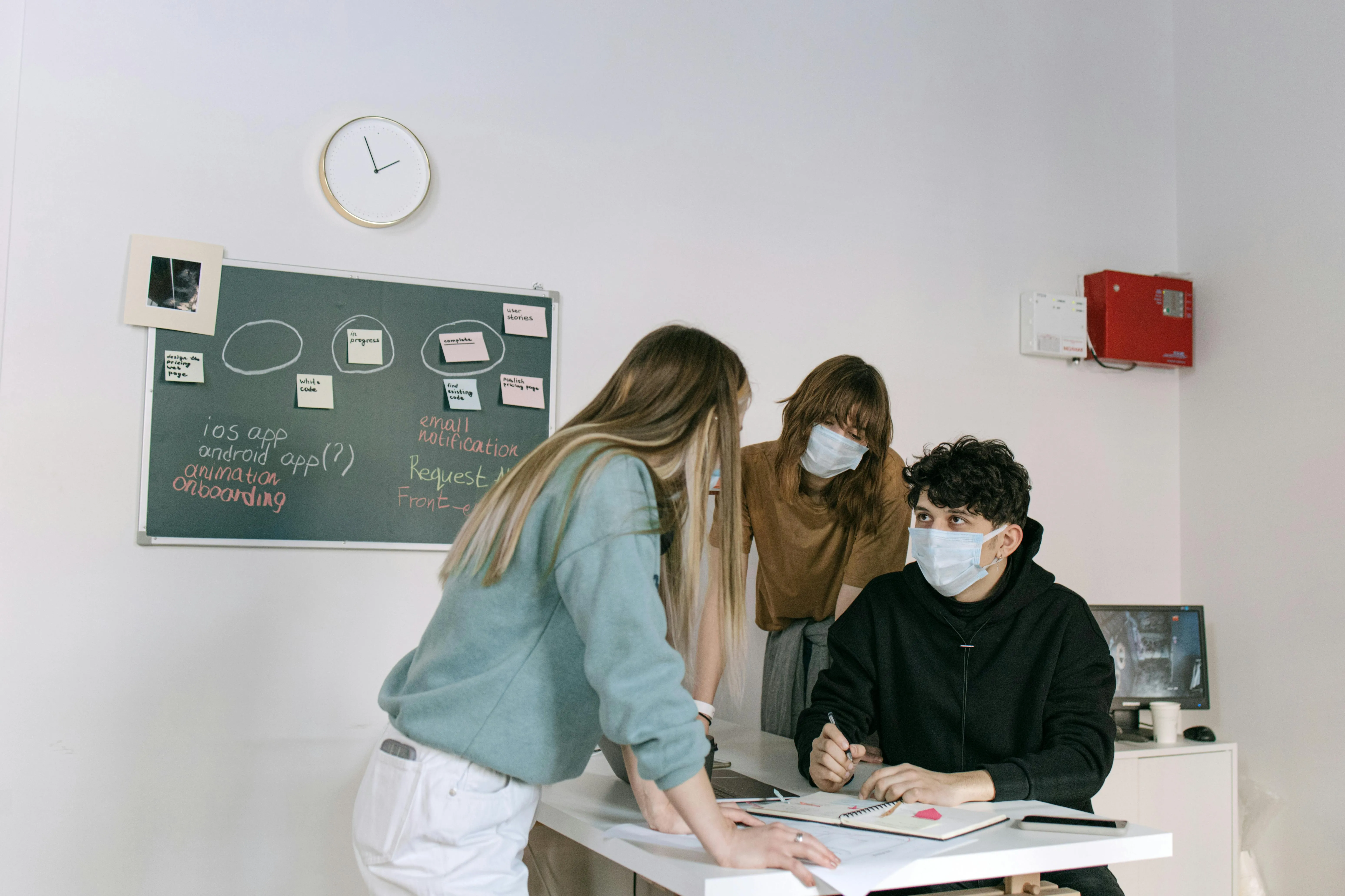 #FotoDescrição Três jovens usando máscaras faciais, reunidos em frente a um quadro com anotações de um projeto digital, representando colaboração e inovação em um ambiente educacional ou criativo.