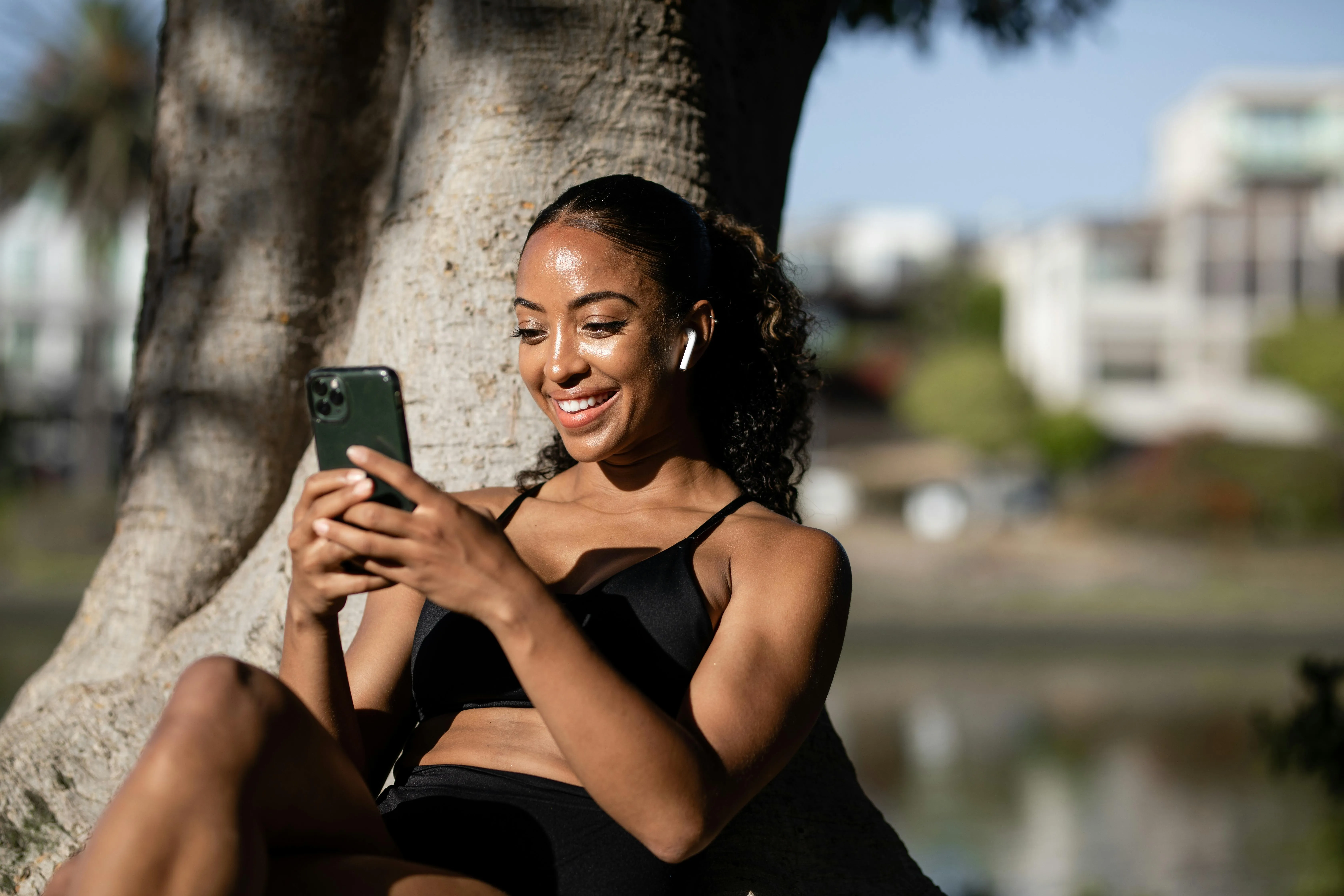 #FotoDescrição: Mulher sorridente, sentada ao ar livre encostada em uma árvore, usando fones de ouvido sem fio enquanto mexe no celular.