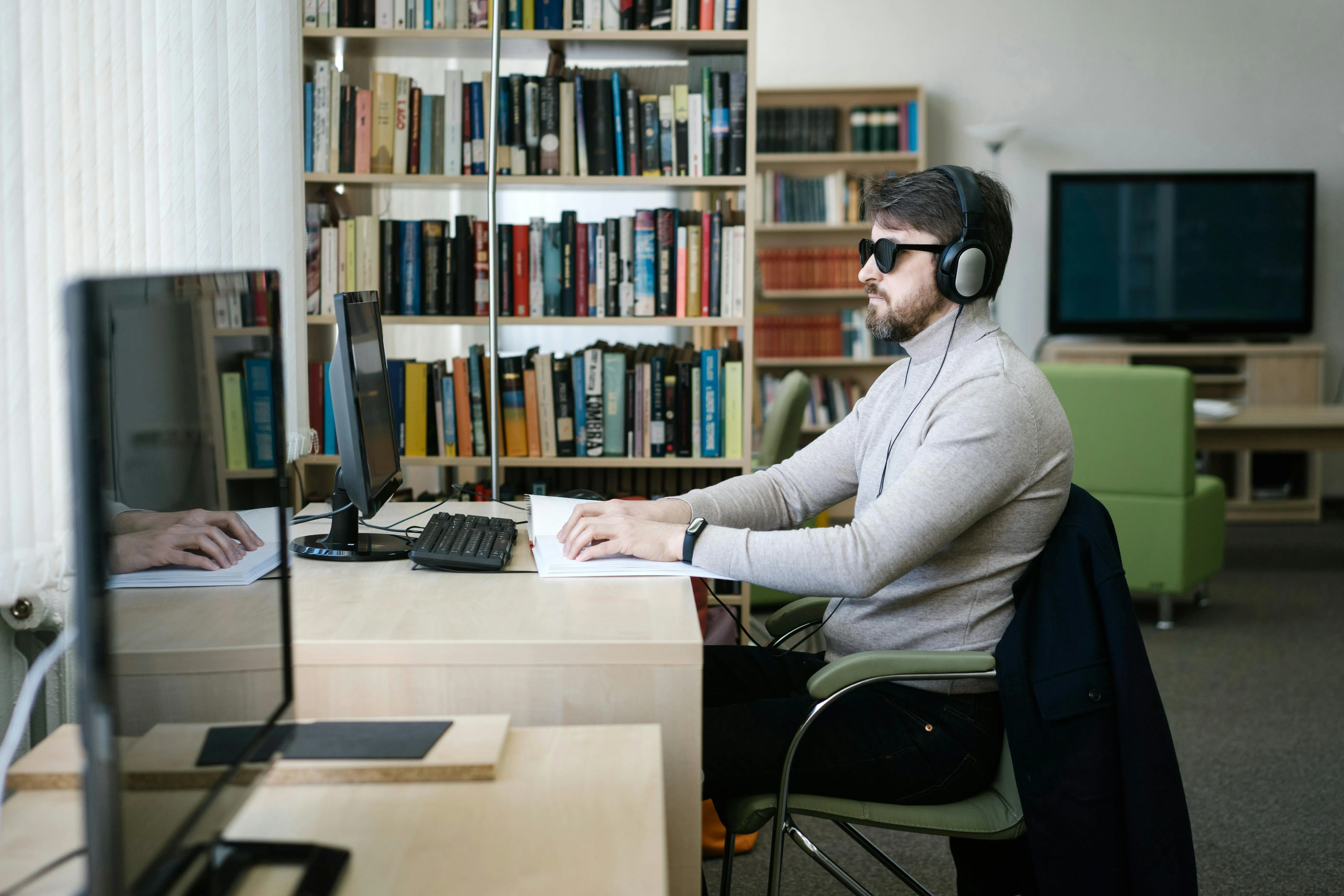Homem em escritório com computadores e livros, usando headset e óculos escuros.