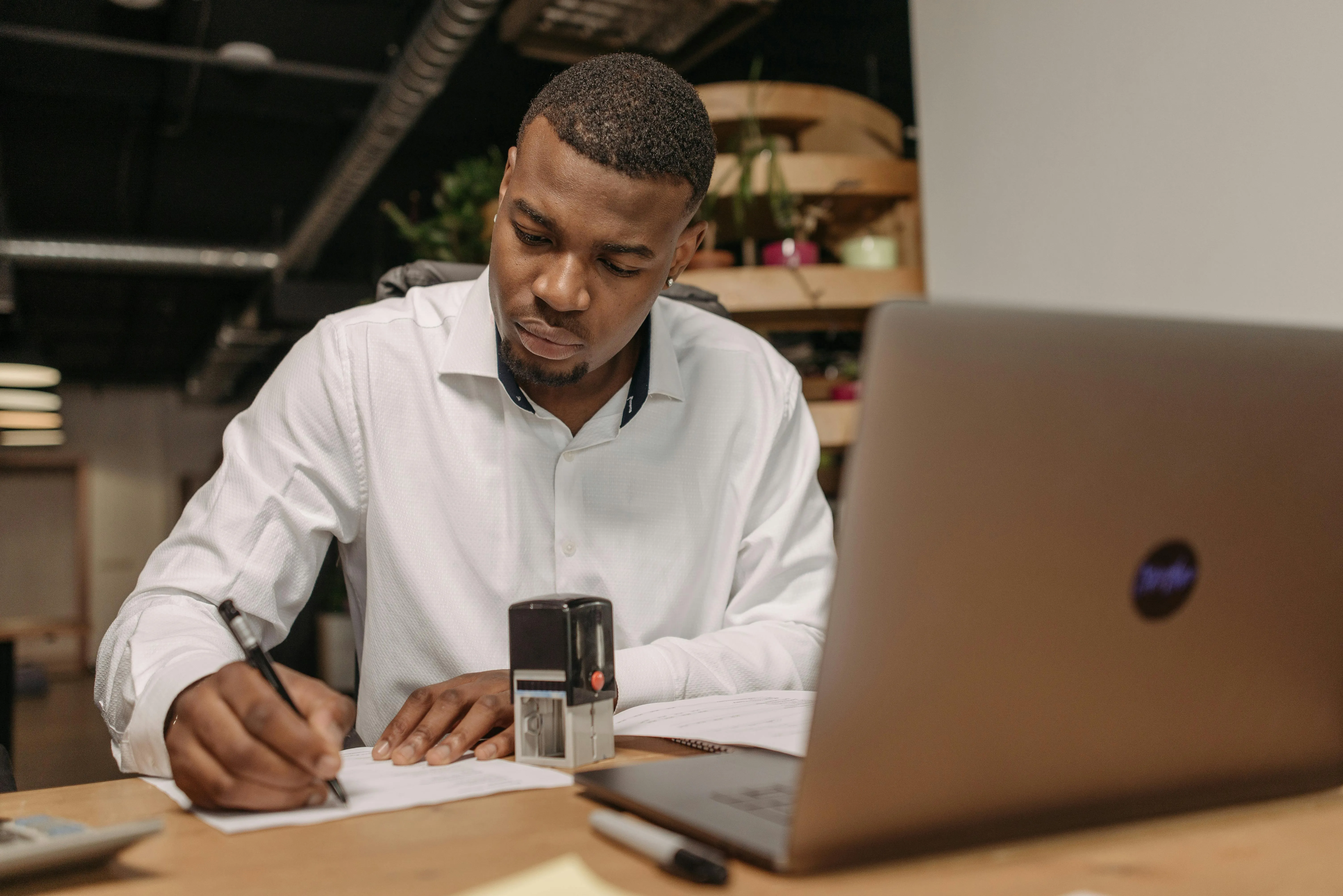 #FotoDescrição Homem escrevendo e carimbando documentos em um escritório, com laptop aberto à frente, simbolizando execução prática de estratégias e organização profissional.