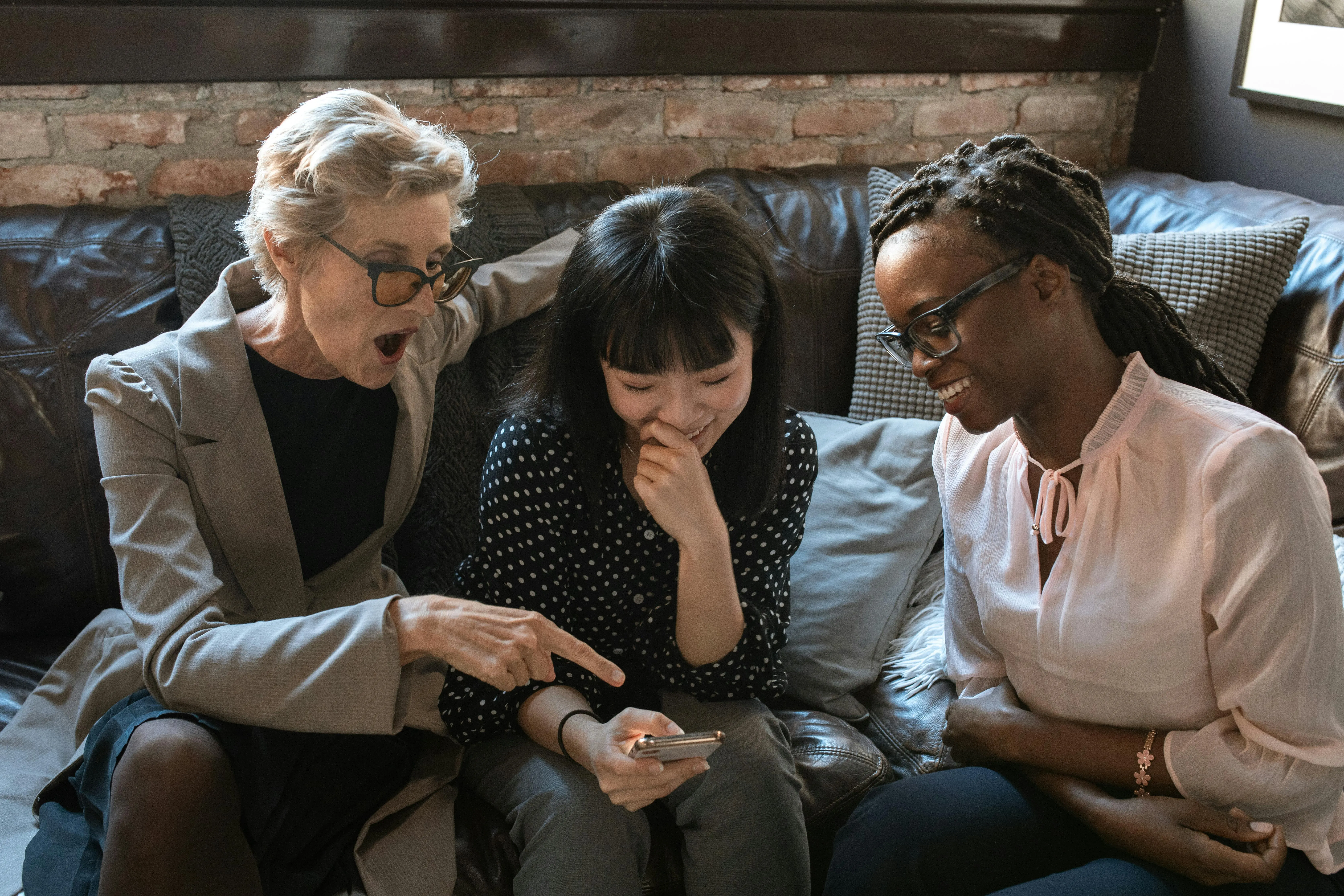 #FotoDescrição: Três mulheres de diferentes idades e etnias sorrindo enquanto olham para um celular, demonstrando interação e troca de ideias.