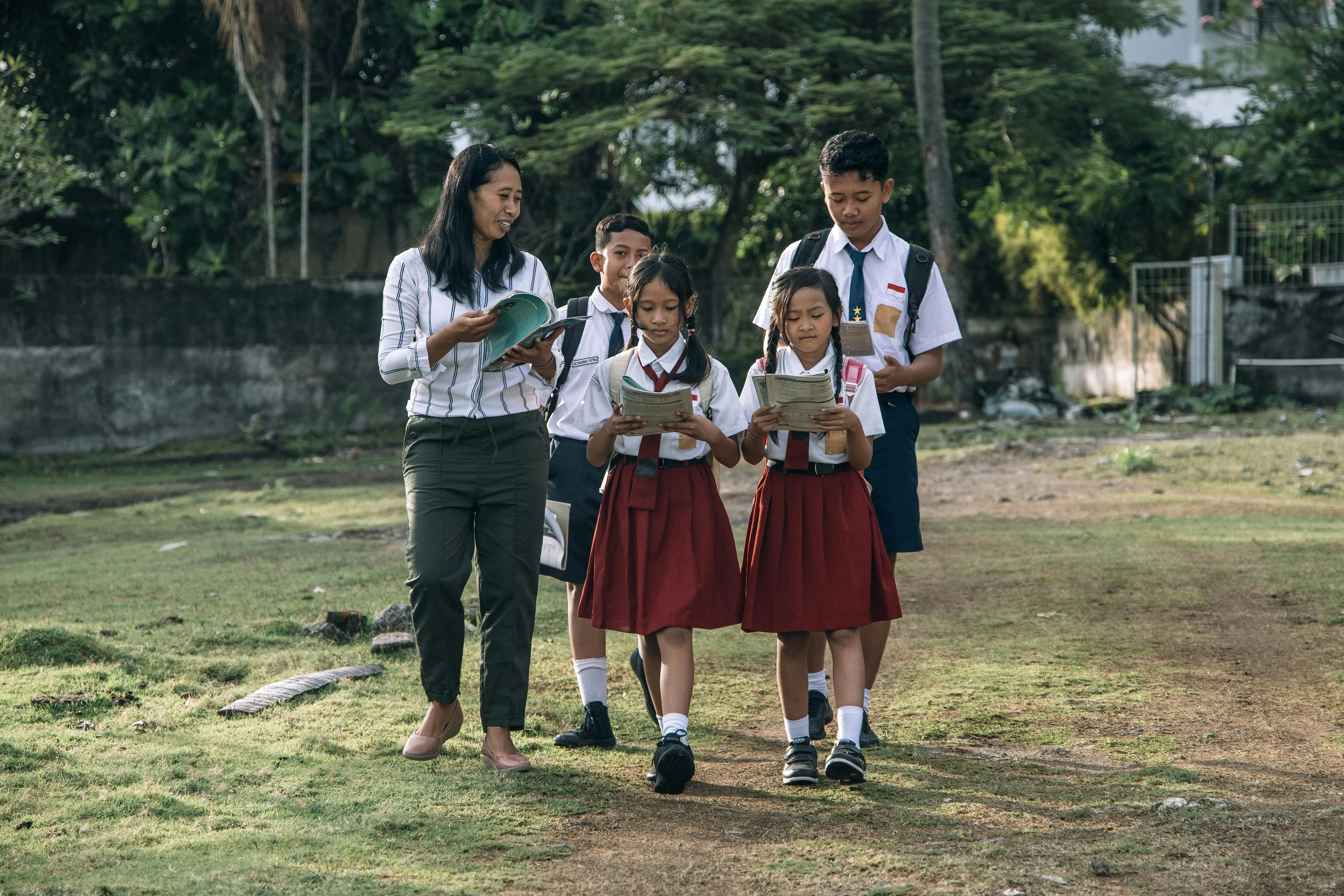 Professora caminha com alunos uniformizados lendo livros ao ar livre.