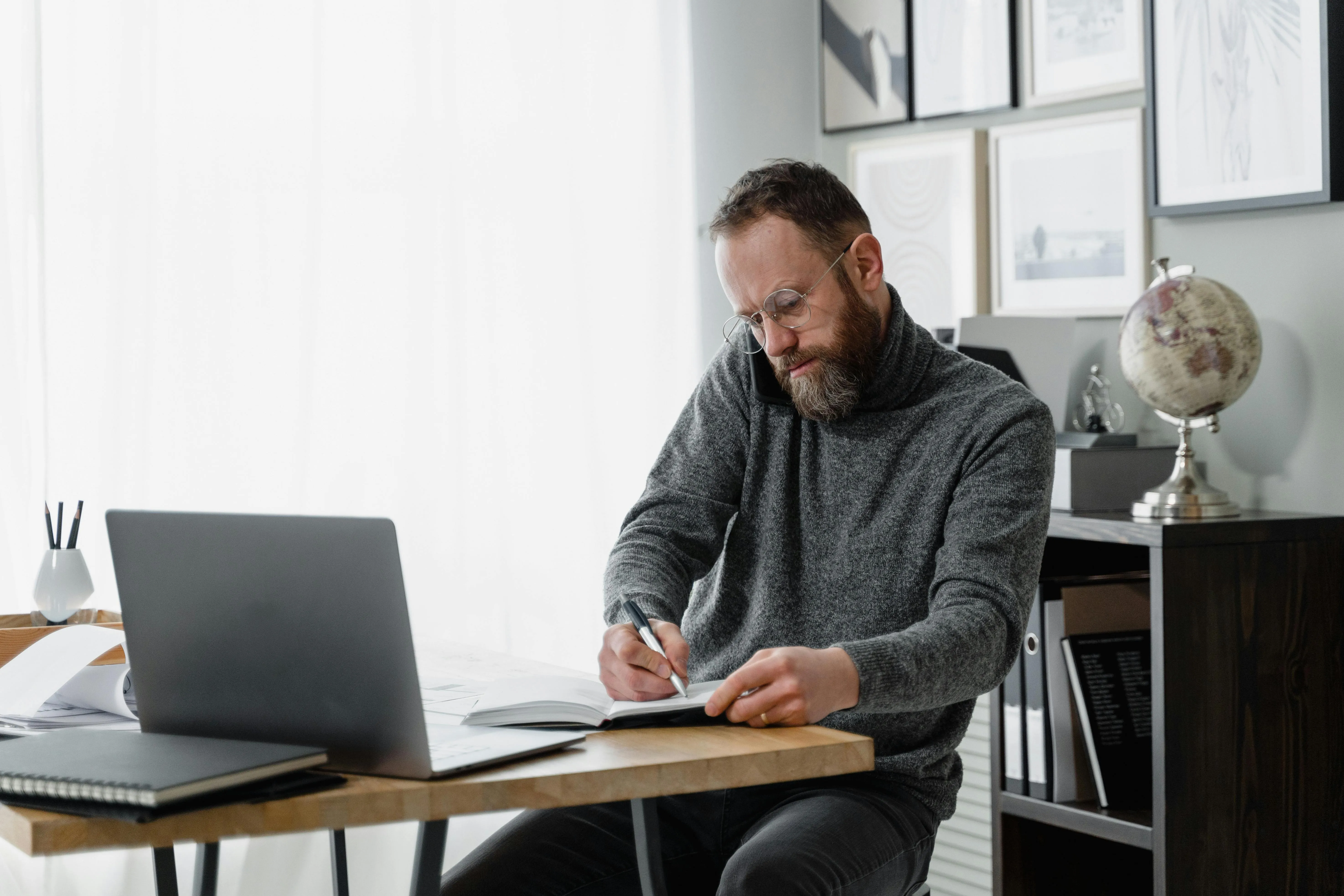 #FotoDescrição Homem com expressão concentrada escrevendo em um caderno enquanto trabalha em um escritório em casa, com notebook à frente e ambiente decorado, simbolizando planejamento e autonomia do consumidor 4.0.