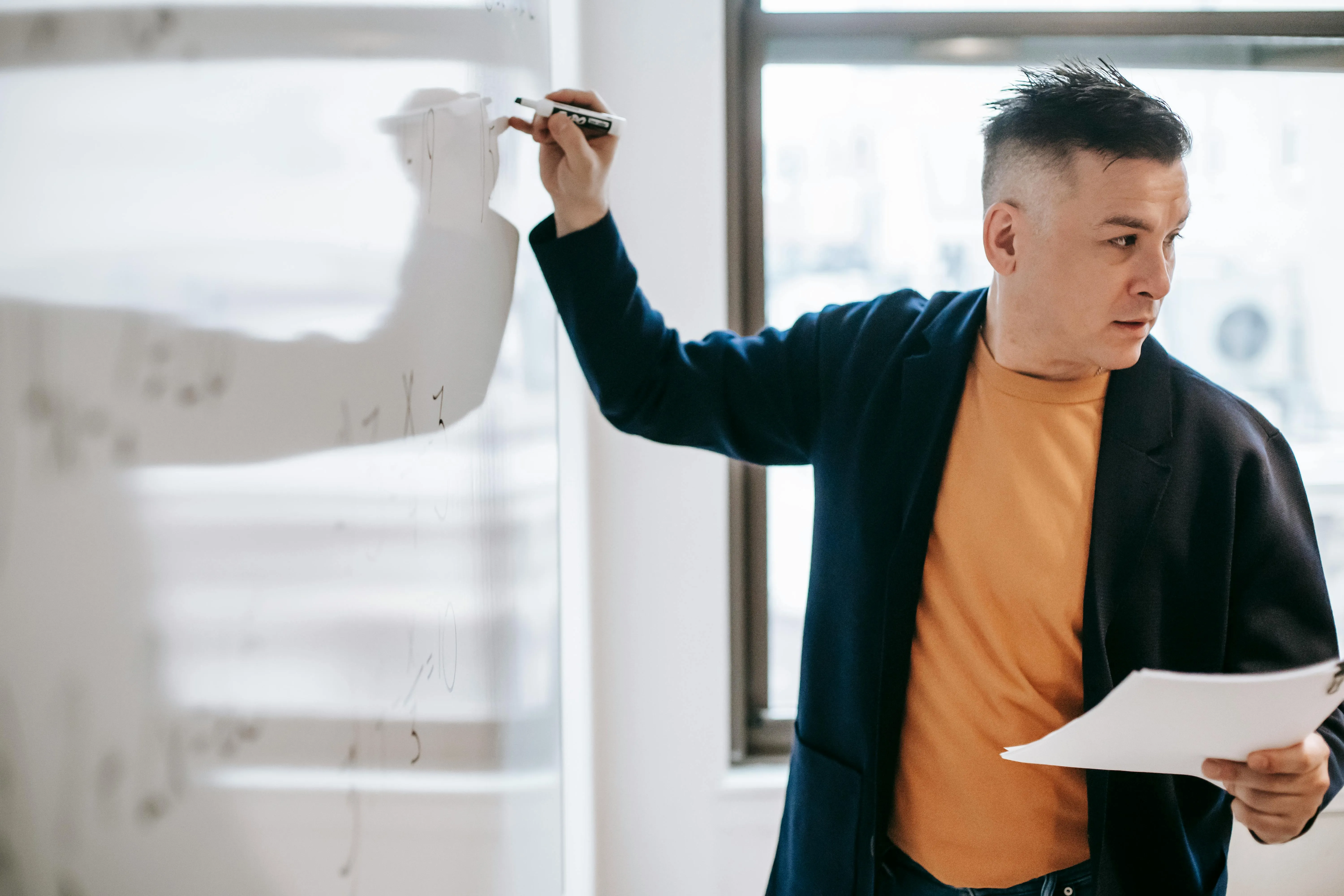 #FotoDescrição: Homem escrevendo em um quadro branco durante uma explicação em sala de aula ou ambiente corporativo.