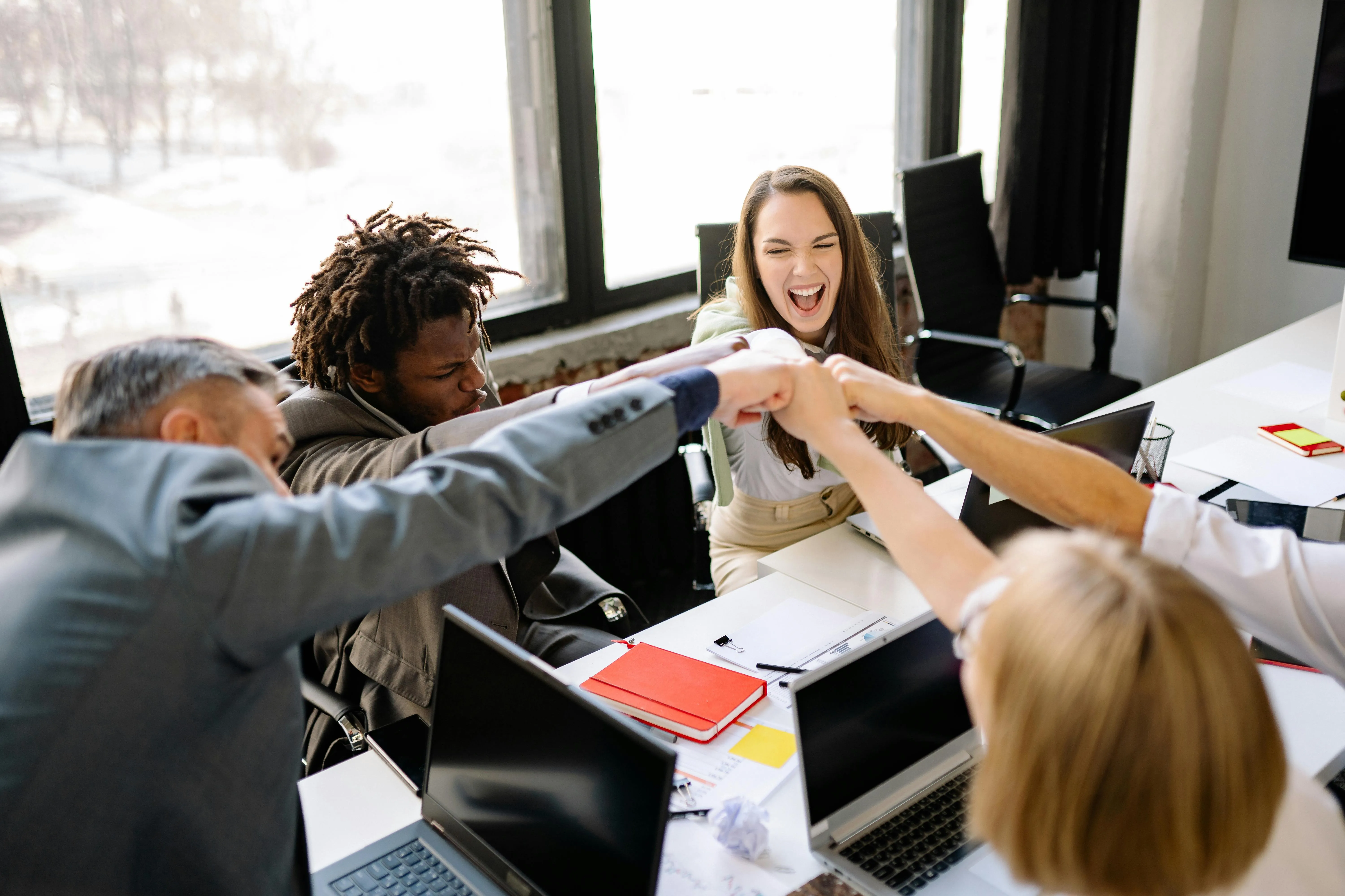 #FotoDescrição: Equipe diversa em sala de reunião fazendo gesto de união com punhos juntos sobre a mesa, sorrindo e celebrando em ambiente descontraído.