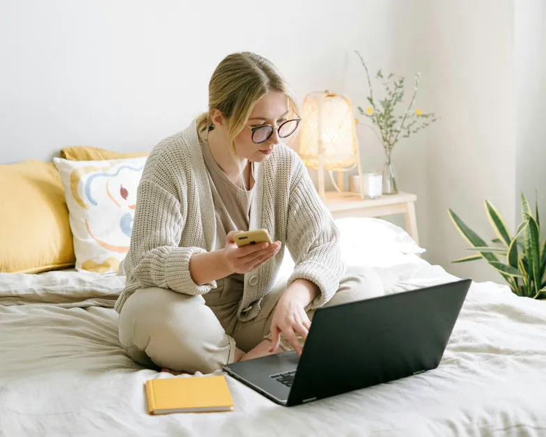 #FotoDescrição: Mulher loira usando um notebook e celular sobre a cama, com caderno amarelo ao lado e decoração minimalista no ambiente.