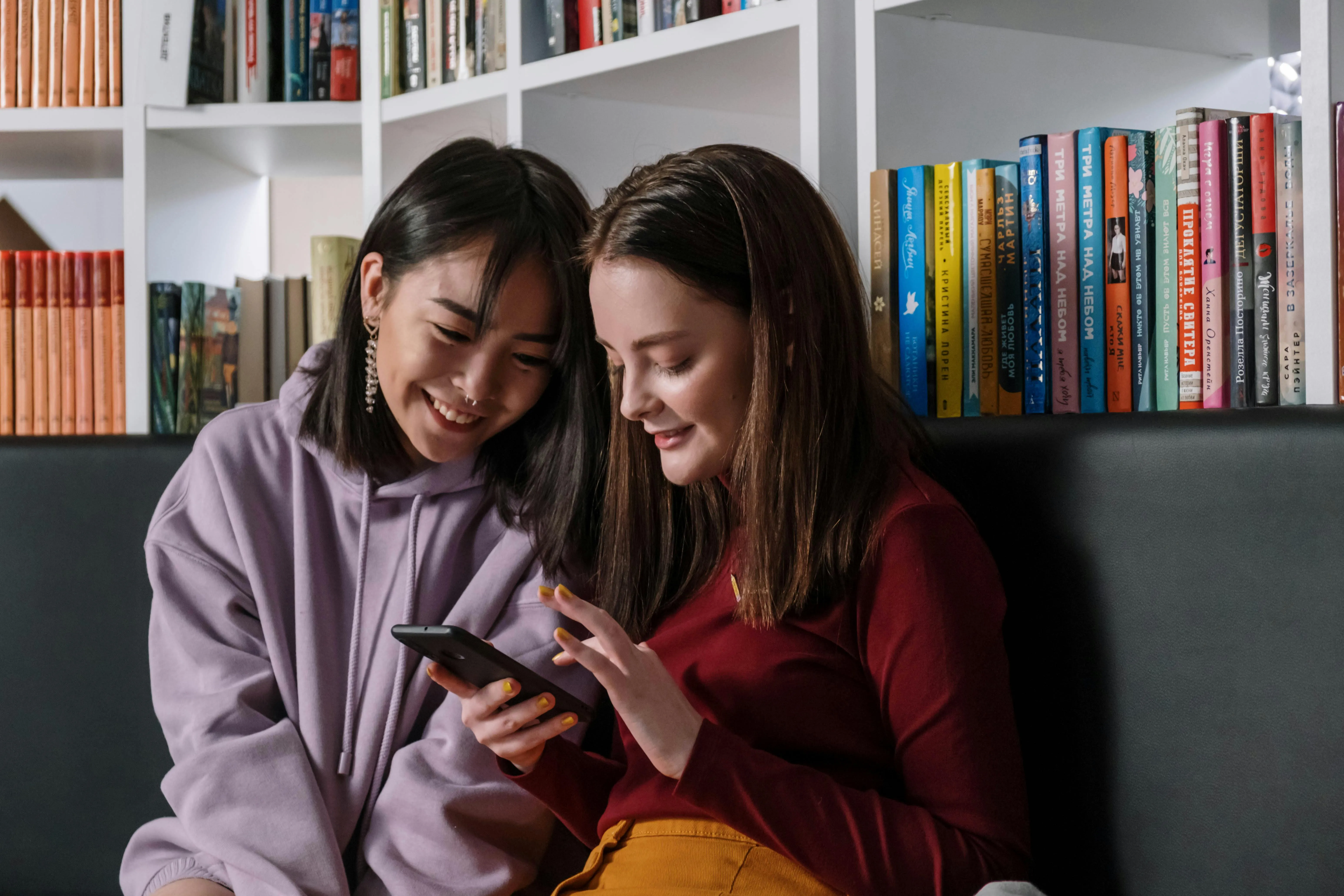 #FotoDescrição: Duas jovens sentadas em uma biblioteca olhando e sorrindo para a tela de um celular.