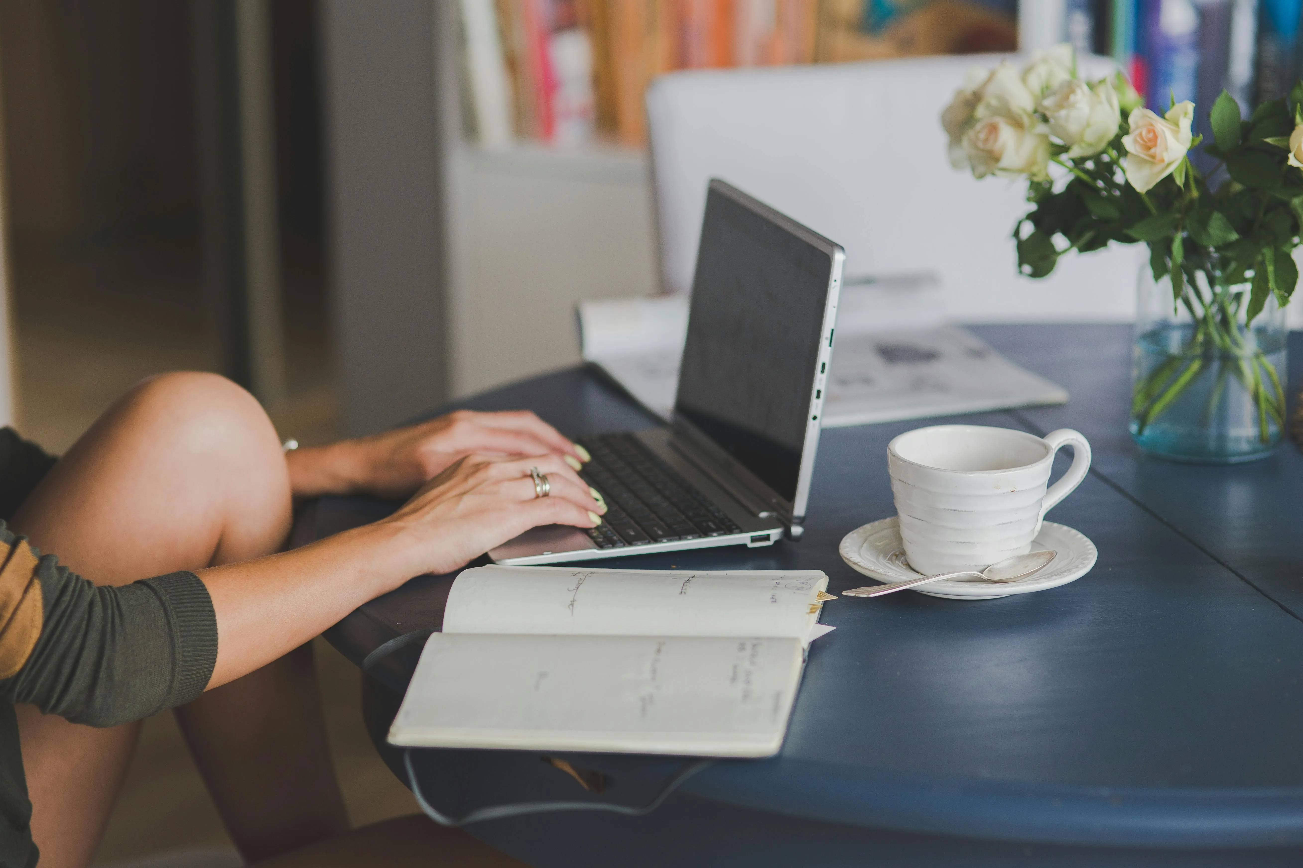 Mulher digitando em laptop com caderno e xícara de café na mesa.