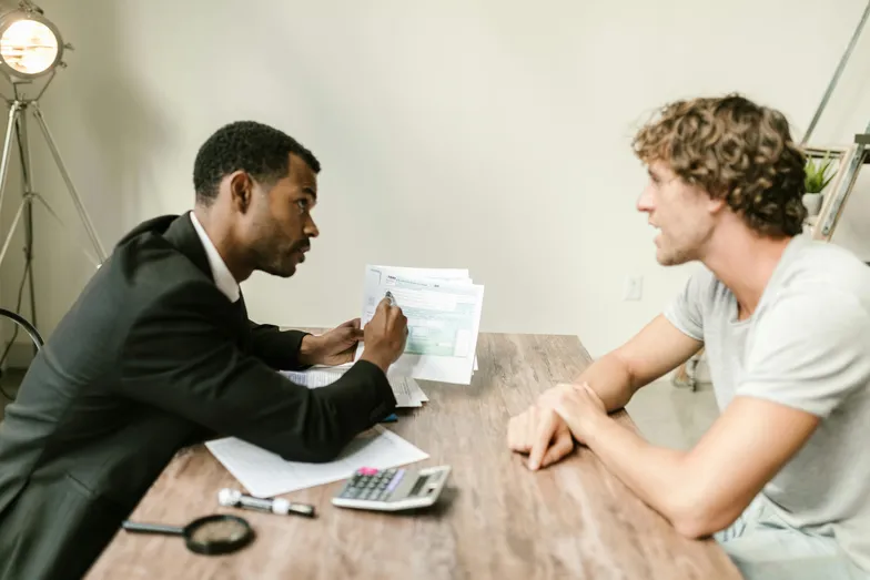 #FotoDescrição: Homem de terno explicando documentos para outro homem de camiseta cinza em uma mesa de madeira, com calculadora e papéis ao redor.