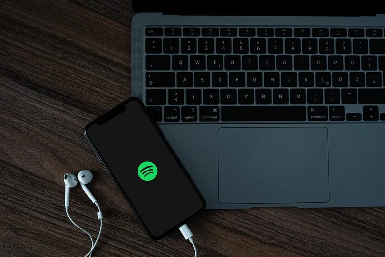 #FotoDescrição: Smartphone com o logo do Spotify na tela, conectado a fones de ouvido e posicionado sobre um notebook em uma mesa de madeira.