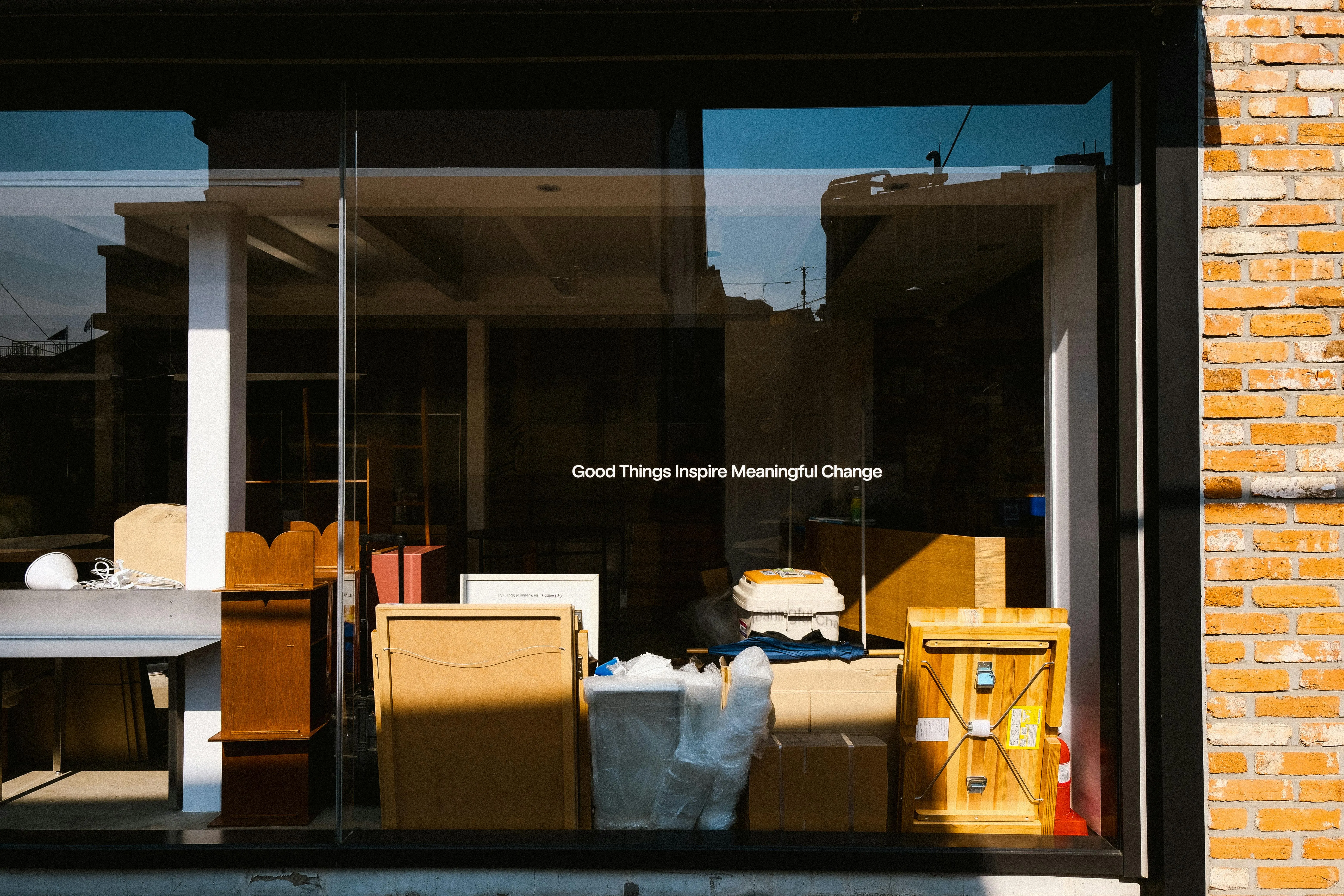 #FotoDescrição Vitrine de uma loja com móveis e objetos diversos à mostra, refletindo a luz do dia e trazendo a frase “Good Things Inspire Meaningful Change” no vidro, simbolizando transformação e criatividade no setor comercial ou imobiliário.