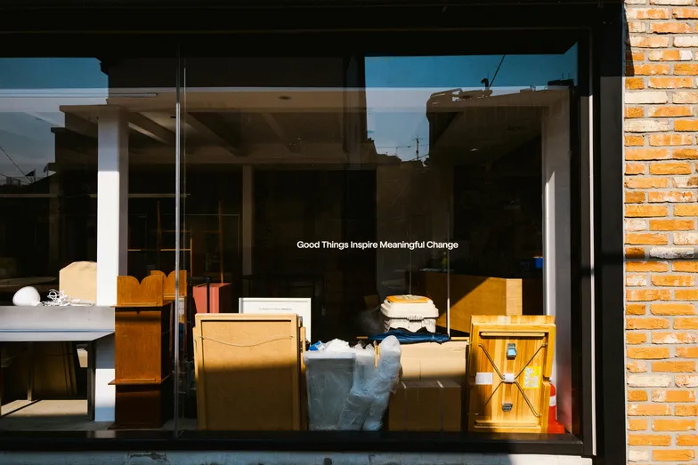 #FotoDescrição Vitrine de uma loja com móveis e objetos diversos à mostra, refletindo a luz do dia e trazendo a frase “Good Things Inspire Meaningful Change” no vidro, simbolizando transformação e criatividade no setor comercial ou imobiliário.