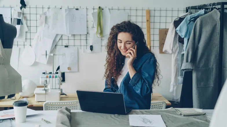 Designer de moda trabalha no laptop e fala ao telefone no ateliê