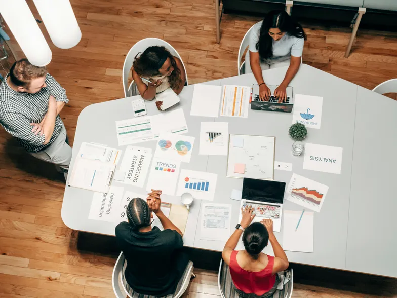 Pessoas reunidas em mesa de trabalho com gráficos e laptops.